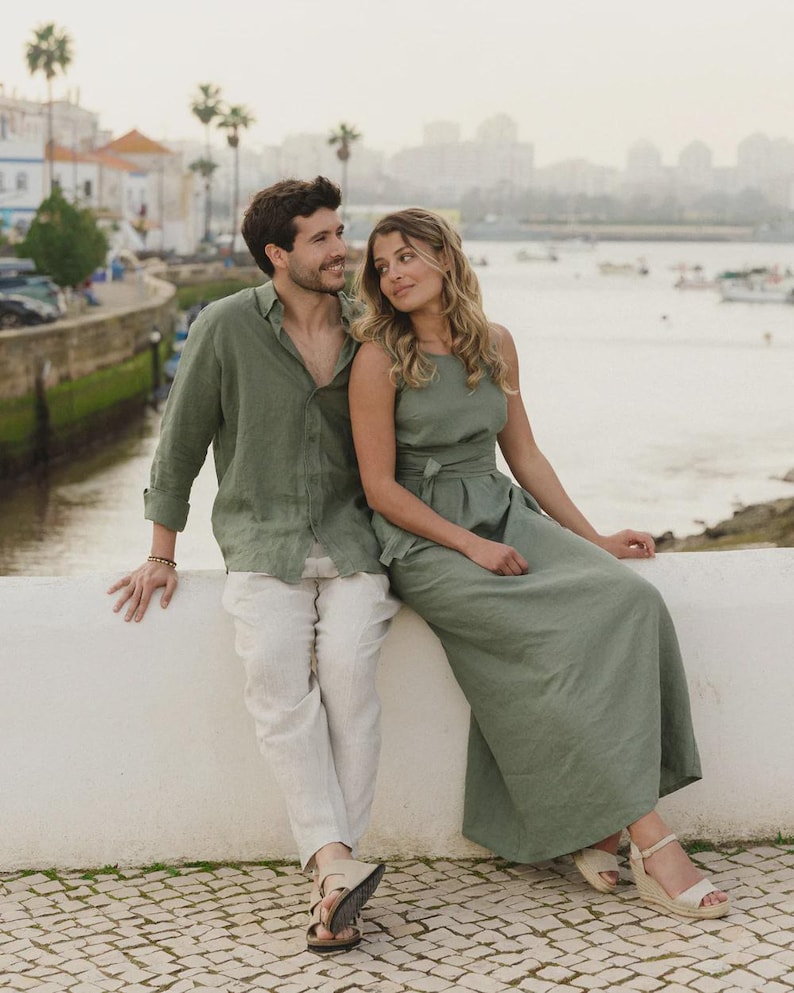 May include: An olive green long-sleeved button-down shirt, light beige trousers, and an olive green sleeveless maxi dress with a tie-waist detail. The clothing is worn with beige sandals and a wedge-heeled sandal, showcasing a coordinated set of apparel.