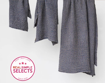 May include: Three dark blue waffle-weave towels of varying sizes hanging from a wooden rod. A circular pink badge in the lower left corner features the text SHOP BUY LOVE, three stars, REAL SIMPLE SELECTS, and SINCE 2000.