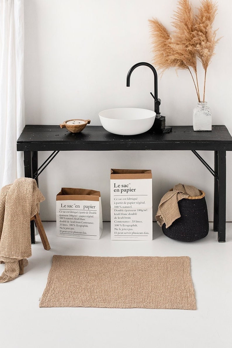 May include: Two brown paper bags with handles, each with the text "Le sac en papier" and information about the bag's material and capacity. The bags are sitting on a black table with a white ceramic sink and a vase with dried pampas grass.