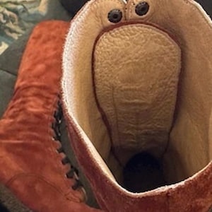 Puede incluir: Primer plano de un par de botas de ante marr&oacute;n rojizo. El interior es de color beige claro con una plantilla texturizada. El borde superior de la bota tiene dos remaches oscuros. Las botas tienen cordones.