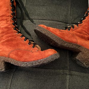 Puede incluir: Un par de botas de ante naranja con cordones negros. Las botas tienen punta redonda, tac&oacute;n bajo y suela texturizada. Las botas son hasta la rodilla y se atan con cordones en la parte delantera.
