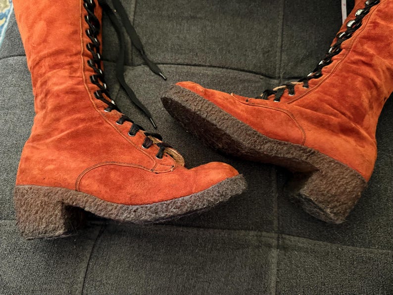 Puede incluir: Un par de botas de ante naranja con cordones negros. Las botas tienen punta redonda, tac&oacute;n bajo y suela texturizada. Las botas son hasta la rodilla y se atan con cordones en la parte delantera.