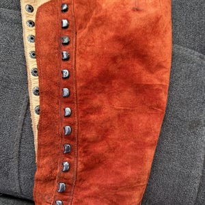 Puede incluir: Primer plano de una bota alta de ante color &oacute;xido. La bota presenta una fila de tachuelas plateadas en el lateral y un panel de cuero color crema con ojales para cordones. El ante tiene un aspecto texturizado.