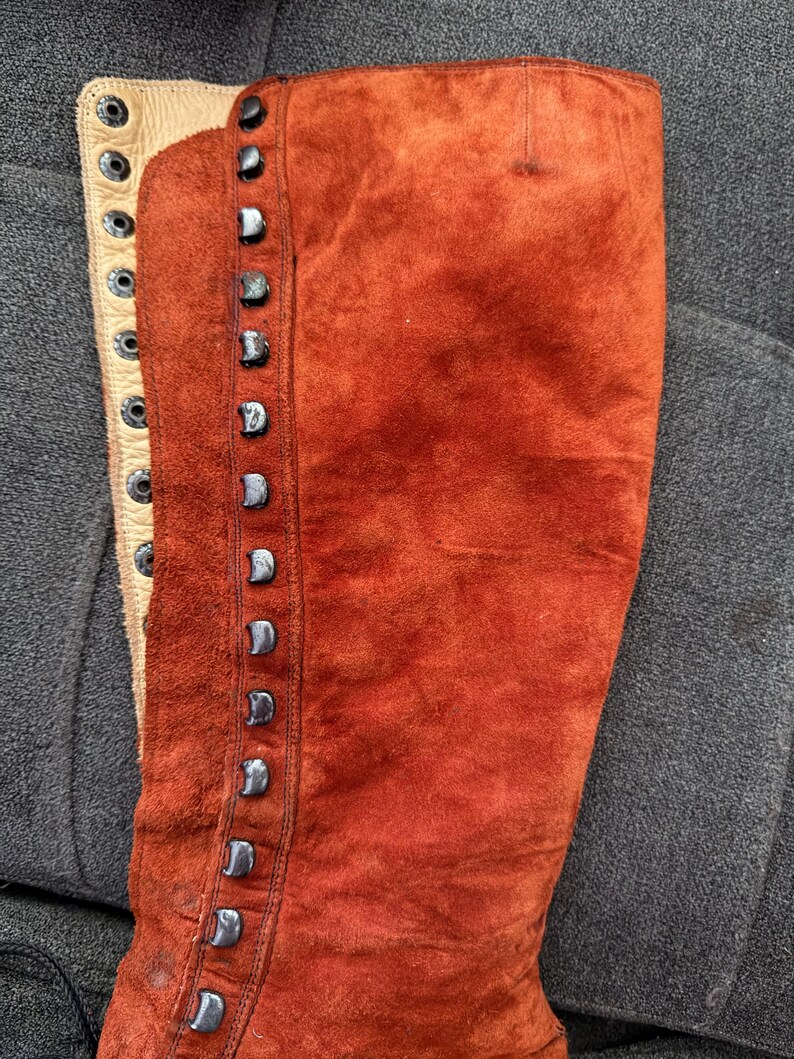 Puede incluir: Primer plano de una bota alta de ante color &oacute;xido. La bota presenta una fila de tachuelas plateadas en el lateral y un panel de cuero color crema con ojales para cordones. El ante tiene un aspecto texturizado.