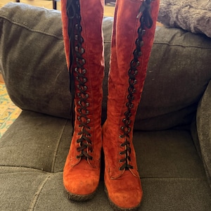 Puede incluir: Un par de botas altas de ante naranja con cordones negros. Las botas tienen punta redonda y suela gruesa. Los cordones suben por la parte delantera de las botas y est&aacute;n atados con un lazo en la parte superior. Las botas est&aacute;n sobre un sof&aacute; gris.