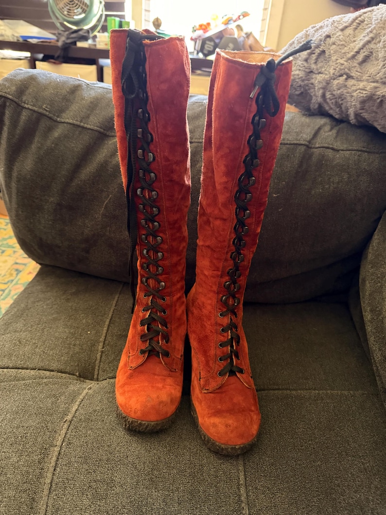 Puede incluir: Un par de botas altas de ante naranja con cordones negros. Las botas tienen punta redonda y suela gruesa. Los cordones suben por la parte delantera de las botas y est&aacute;n atados con un lazo en la parte superior. Las botas est&aacute;n sobre un sof&aacute; gris.