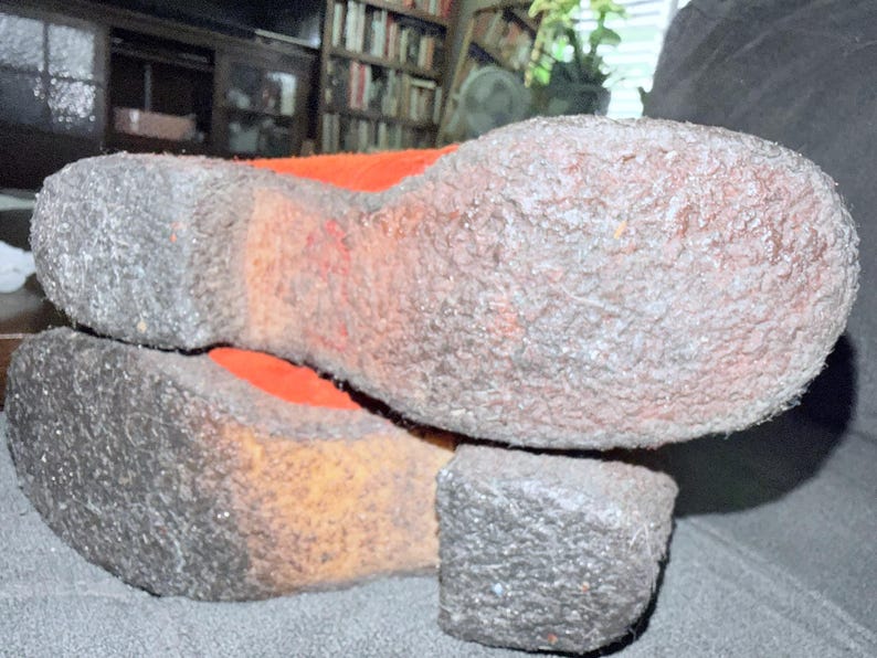 Puede incluir: Primer plano de las suelas de dos zapatos. Las suelas son de color gris claro texturizado con un tal&oacute;n y una puntera gris oscuro. El interior del zapato es naranja. Los zapatos descansan sobre una superficie gris oscuro.