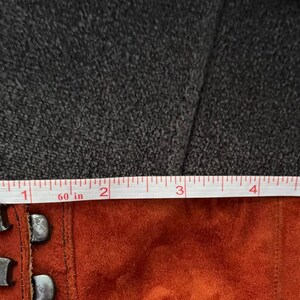 Puede incluir: Primer plano de un material de gamuza color &oacute;xido con herrajes met&aacute;licos. Una cinta m&eacute;trica muestra una longitud de aproximadamente 15 cm. El fondo es una superficie oscura y texturizada.