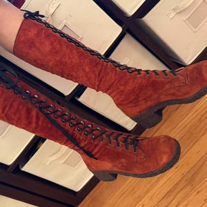 Puede incluir: Un par de botas altas de ante color &oacute;xido con cordones negros. Las botas tienen una puntera redondeada y un tac&oacute;n peque&ntilde;o y oscuro. Los cordones suben por la parte delantera y est&aacute;n atados en la parte superior.