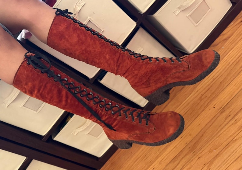 Puede incluir: Un par de botas altas de ante color &oacute;xido con cordones negros. Las botas tienen una puntera redondeada y un tac&oacute;n peque&ntilde;o y oscuro. Los cordones suben por la parte delantera y est&aacute;n atados en la parte superior.