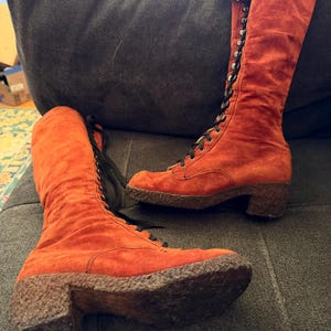 Puede incluir: Un par de botas altas de ante naranja con cordones negros. Las botas tienen una suela texturizada y un tac&oacute;n peque&ntilde;o. Las botas son hasta la rodilla y parecen ser vintage.