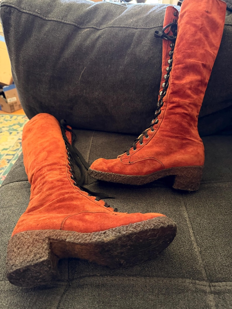 Puede incluir: Un par de botas altas de ante naranja con cordones negros. Las botas tienen una suela texturizada y un tac&oacute;n peque&ntilde;o. Las botas son hasta la rodilla y parecen ser vintage.
