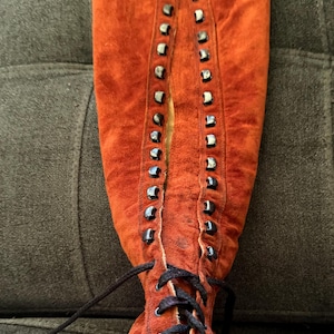 Puede incluir: Una bota alta de ante color &oacute;xido con cordones en la parte delantera. La bota presenta ojales met&aacute;licos y un cord&oacute;n negro. El ante tiene un aspecto ligeramente desgastado. La bota es de estilo vintage.