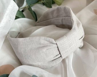 May include: A gray linen headband with a knotted detail. The headband is on a white background with green leaves.