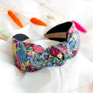 Puede incluir: Una diadema con estampado floral con un diseño anudado. La diadema presenta un colorido patrón de flores en tonos rosa, azul, amarillo y verde. El interior es negro y beige.