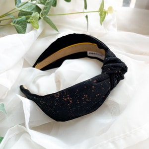 May include: A wide black fabric headband featuring a knotted top detail and a speckled pattern of metallic copper-colored dots. The interior of the headband is lined with a tan material and includes a white label with the text "nine designs".