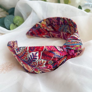 May include: A knotted fabric headband featuring a vibrant, multi-colored botanical print. The pattern displays intricate leaves and floral motifs in shades of deep red, bright pink, blue, yellow, and white, arranged in a dense, repeating design.