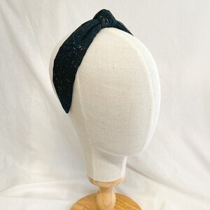 May include: A black fabric headband featuring a knotted top detail and a speckled pattern of tiny white and copper-colored dots. The headband is displayed on a neutral-toned mannequin head, showcasing the fabric texture and the overall shape.