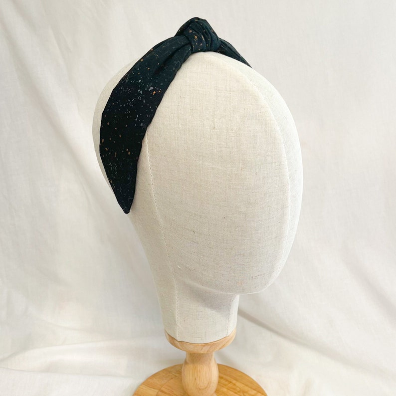 May include: A black fabric headband featuring a knotted top detail and a speckled pattern of tiny white and copper-colored dots. The headband is displayed on a neutral-toned mannequin head, showcasing the fabric texture and the overall shape.