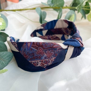 May include: A blue, white, and burgundy patterned knotted headband. The headband is made of fabric and has a paisley design.