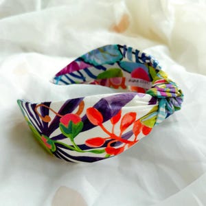 May include: A patterned fabric headband with a knotted design. The headband displays a white base adorned with a floral and striped pattern in purple, red, green, and blue. The headband is approximately 2.5 cm wide.
