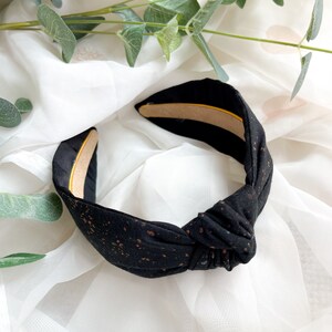 May include: A black knotted headband featuring a subtle copper-colored speckled pattern throughout the fabric. The interior of the headband is lined with a smooth, tan-colored material, providing a contrast to the dark exterior fabric.