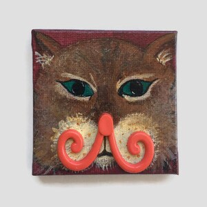 Puede incluir: Una pintura de la cara de un gato con un bigote rojo y rizado. El gato tiene pelaje marrón, ojos verdes y una nariz blanca.