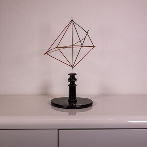Può includere: Una scultura geometrica caratterizzata da una struttura in filo metallico con aste alternate di colore nero e rame. La forma angolare e sfaccettata è montata su una solida base a piedistallo cilindrica di colore nero.