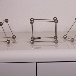 May include: Three silver-toned wire sculptures, each consisting of eight small spheres connected by thin metal rods to form geometric cube or rhomboid structures. The centre sculpture features a small black rectangular tag with white text.