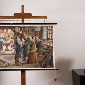 May include: A vintage educational wall chart depicting a saddlery workshop with craftspeople working on leather goods, mounted on a wooden hanging frame. Beside it sits a white ceramic sculpture of a human face with an open mouth on a black base.
