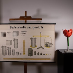 May include: A vintage educational wall chart titled Dezimalmaße und-gewichte, displaying illustrations of metric units, containers, weights, and a balance scale. Beside it stands a decorative red tulip sculpture with a green stem on a base.