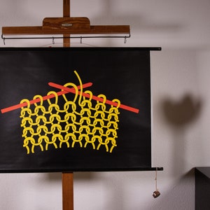 Könnte beinhalten: Ein schwarzes Wandplakat mit einer grafischen Darstellung von gelben Garnschleifen, die auf zwei horizontalen roten Stricknadeln gearbeitet werden. Das Plakat ist auf einem Holzrahmen mit hängenden Gewichten an den unteren Ecken montiert.