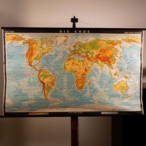 Könnte beinhalten: Eine vintage Wandkarte zum Herunterziehen mit dem Titel DIE ERDE, die eine physische Weltkarte mit topographischer Schattierung in Grün-, Gelb- und Brauntönen zeigt. Die Karte ist an schwarzen Holzstäben mit einer Aufhängeschnur befestigt und zeigt den Maßstab 1:24 000 000.