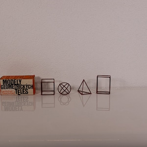 Könnte beinhalten: Ein Set aus braunen Draht-Geometriemodellen, bestehend aus einem Würfel, einer Kugel, einer Pyramide und einem Quader, neben einer orangefarbenen Schachtel mit der Aufschrift "MODELY GEOMETRICKÝCH TĚLES DRÁTĚNÉ ŽÁKOVSKÉ".