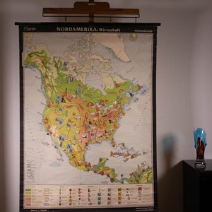 May include: A vintage educational wall map titled NORDAMERIKA: Wirtschaft, featuring a detailed economic overview of North America with various icons representing industries, agriculture, and mining, mounted on a wooden hanging rod system.