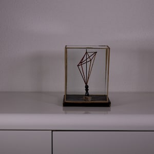 Può includere: Una scultura geometrica composta da sottili bastoncini di legno chiaro disposti a forma di diamante, racchiusa in una teca rettangolare in vetro con base scura. La struttura è sostenuta da un piccolo piedistallo verticale scuro.