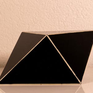 Può includere: Una scultura geometrica nera caratterizzata da molteplici facce triangolari unite insieme. L'oggetto presenta bordi netti e definiti e una finitura superficiale scura e liscia. La struttura è composta da diversi piani interconnessi che creano una forma tridimensionale complessa e angolare con sottili linee chiare visibili lungo le giunture dove i pannelli si incontrano.