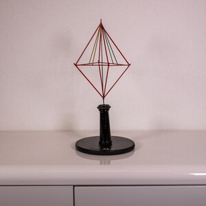 Op de afbeelding: Een geometrisch sculptuur met een rood draadframe in de vorm van een dubbele piramide, ondersteund door een centrale verticale staaf en een ronde zwarte voet. Dunne gekleurde draden verbinden de bovenste top met het middelste frame.