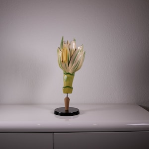 May include: A botanical model of a flower bud featuring cream-colored petals with green stripes, a green stem, and a yellow stamen. The model is mounted on a wooden post attached to a circular black base, displaying a detailed anatomical structure.