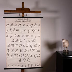 Könnte beinhalten: Eine vintage Lehrtafel mit dem Lateinische Ausgangsschrift Schreibschrift-Alphabet in schwarzer Tinte auf cremefarbenem Hintergrund, aufgehängt an einem Holzrahmen. Die Tafel zeigt Klein- und Großbuchstaben, Satzzeichen und Zahlen.