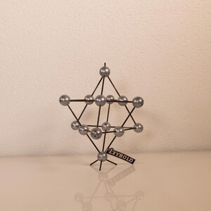 Könnte beinhalten: Ein Leybold Molekülmodell bestehend aus silberfarbenen Kugeln, die durch dünne schwarze Stäbe zu einer geometrischen Gitterstruktur verbunden sind. Das Modell hat eine komplexe, vielzackige Form mit einem kleinen schwarzen Schild mit der Aufschrift LEYBOLD.