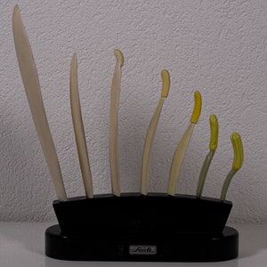 Puede incluir: Un conjunto de siete herramientas de escultura de diversas longitudes y formas, con puntas de color beige claro y amarillo, dispuestas en un soporte negro con la marca "Saidi" impresa en la base frontal. Las herramientas están verticales y ordenadas.