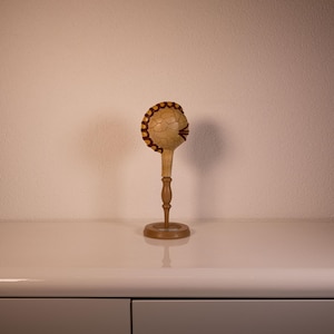 Puede incluir: Maraca de madera hecha a mano con una cabeza redonda y texturizada y un mango largo y delgado. La cabeza presenta un patrón decorativo con detalles en rojo. La maraca se exhibe sobre una superficie clara sobre un fondo neutro.