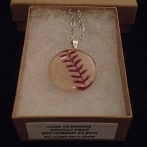 Puede incluir: Un collar de cadena de plata con un colgante de béisbol de vidrio. La pelota de béisbol es blanca con costuras rojas y tiene el texto "CUBS VS BRAVES WRIGLEY FIELD SEPTEMBER 21 2013 BALLHAWK.NET # 183088" en la parte posterior.