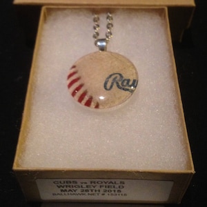 Puede incluir: Un collar de cadena de plata con un colgante redondo de vidrio que presenta una pelota de béisbol con rayas rojas y blancas y la palabra "Rays" en letras azules. El colgante está en una pequeña caja con el texto "CUBS vs ROYALS WRIGLEY FIELD MAY 28TH 2015 BALLHAWK NET # 153115".
