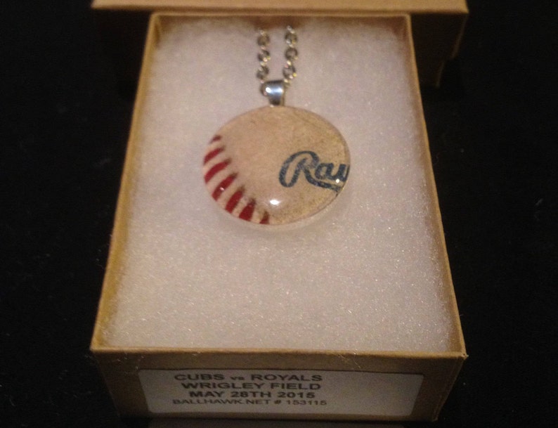 Op de afbeelding: Een zilveren ketting met een ronde glazen hanger met een baseball met rode en witte strepen en het woord "Rays" in blauwe letters. De hanger zit in een klein doosje met de tekst "CUBS vs ROYALS WRIGLEY FIELD MAY 28TH 2015 BALLHAWK NET # 153115".