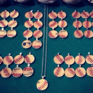 Op de afbeelding: Een zilveren ketting met meerdere baseball-thema hangers. De hangers zijn rond en hebben een baseball-ontwerp met rode stiksels. De ketting wordt getoond op een groene ondergrond met verschillende baseballs.