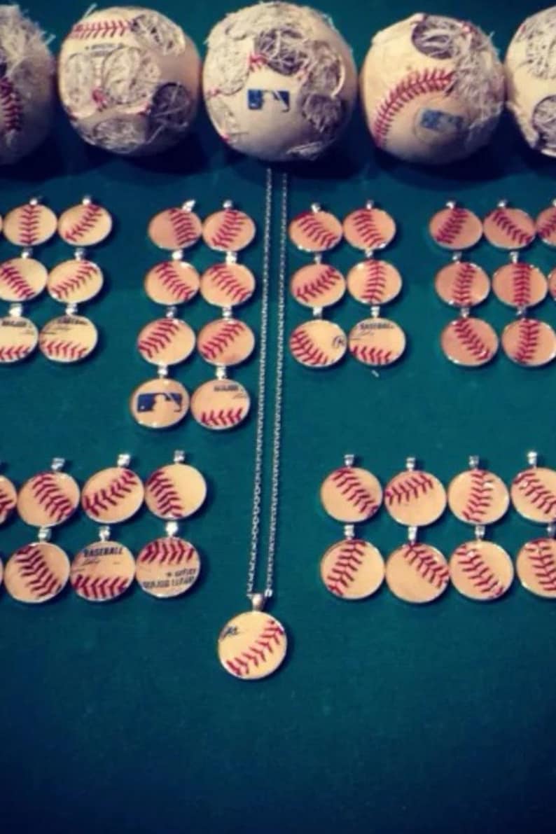 Op de afbeelding: Een zilveren ketting met meerdere baseball-thema hangers. De hangers zijn rond en hebben een baseball-ontwerp met rode stiksels. De ketting wordt getoond op een groene ondergrond met verschillende baseballs.