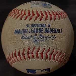 May include: A white baseball with red stitching and the text "OFFICIAL MAJOR LEAGUE BASEBALL" in blue. The baseball also has the signature "Rob Manfred Jr. Commissioner" in blue.