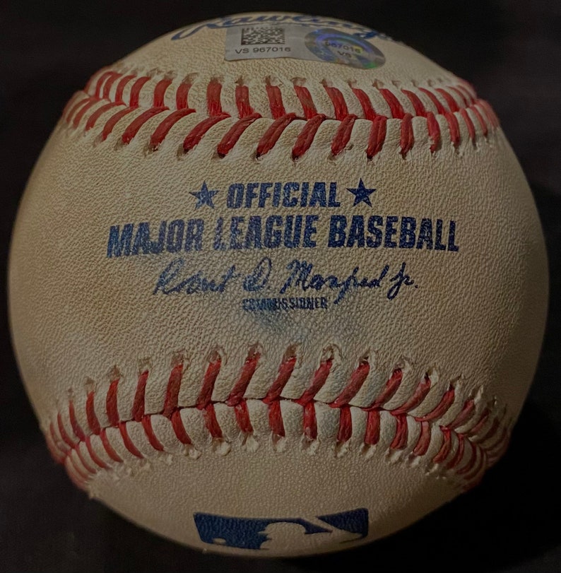 May include: A white baseball with red stitching and the text "OFFICIAL MAJOR LEAGUE BASEBALL" in blue. The baseball also has the signature "Rob Manfred Jr. Commissioner" in blue.